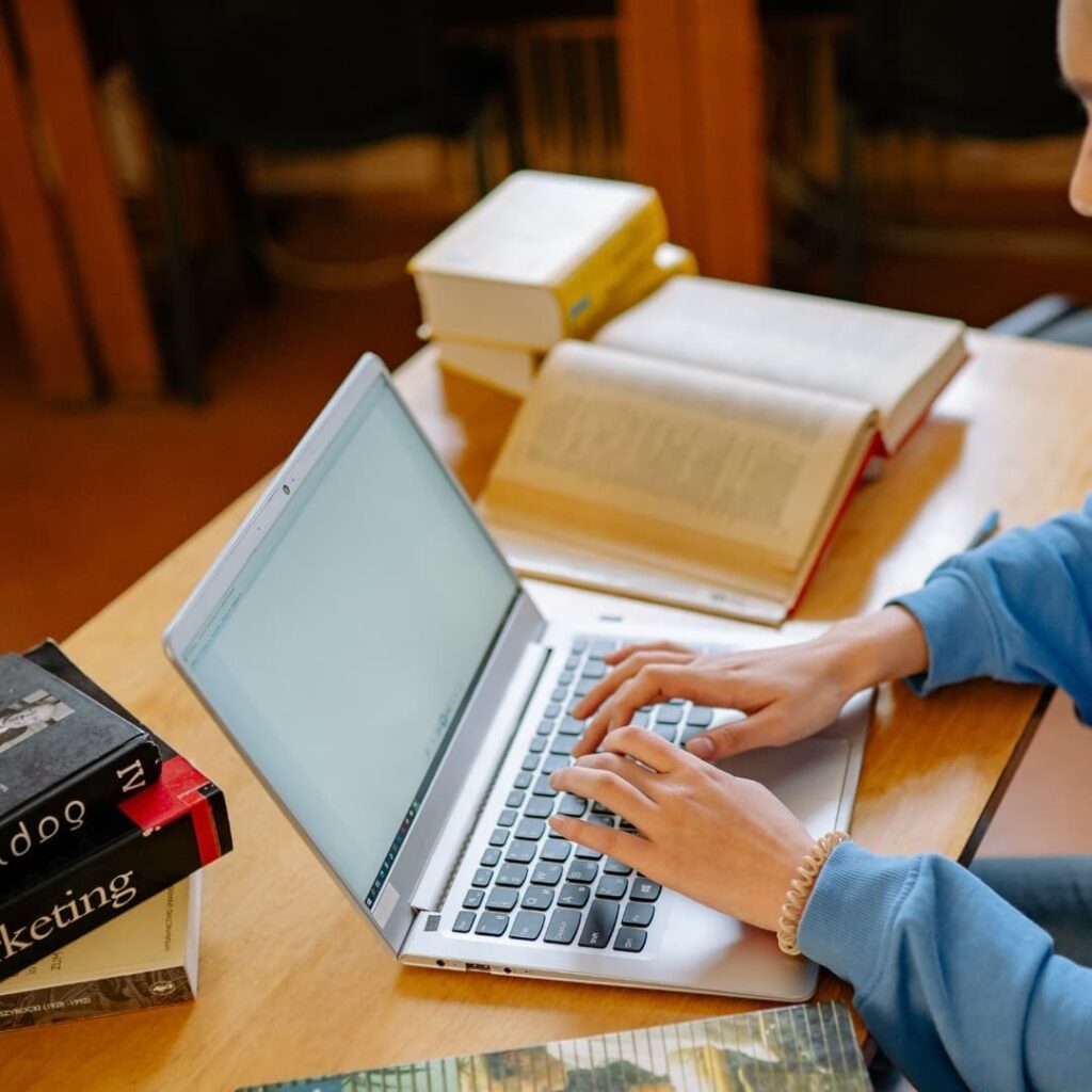 Person typing on a laptop at a desk with books open for studying.