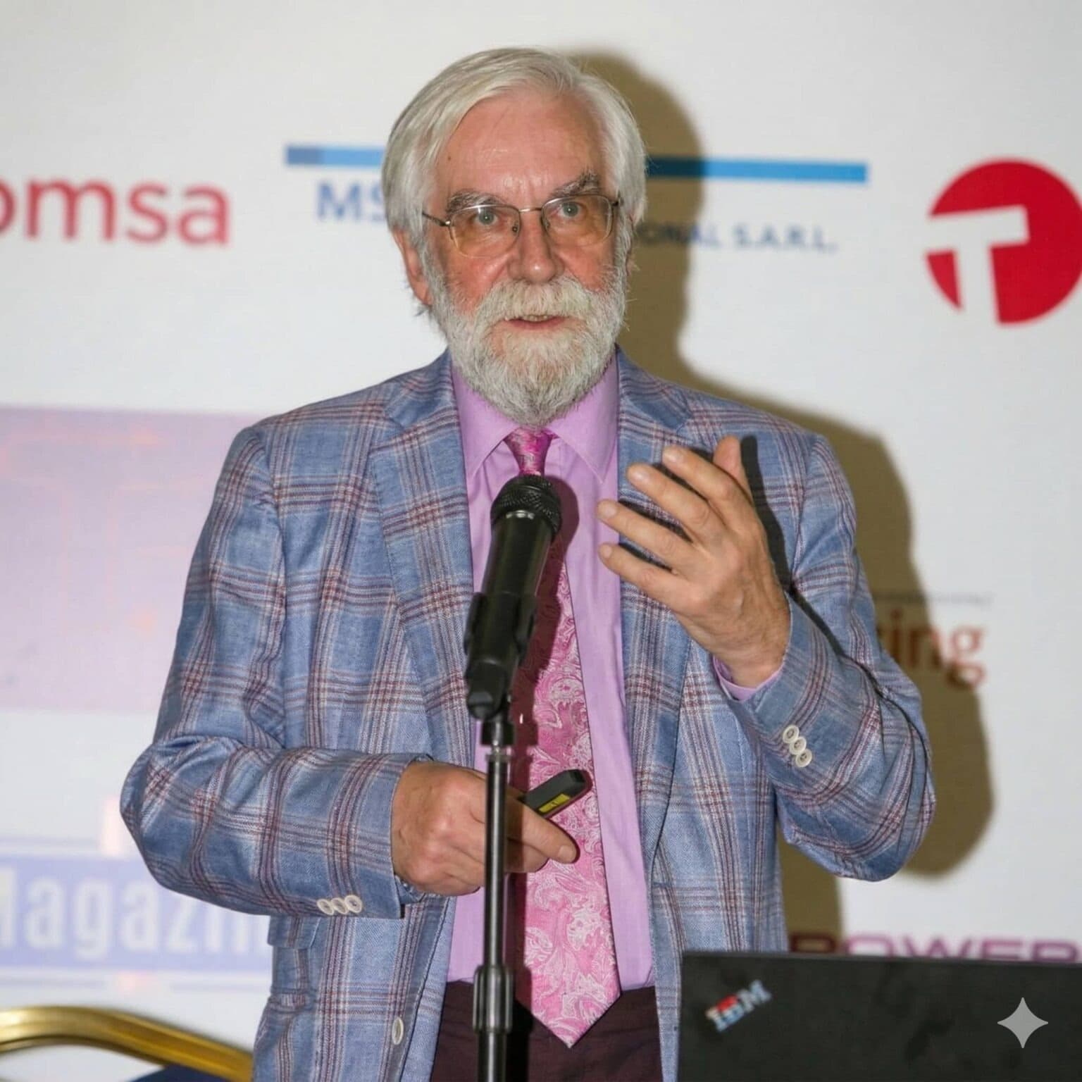 Roy speaking at a conference, wearing glasses, a patterned blue blazer, pink shirt, and tie while holding a microphone and a phone during a presentation.